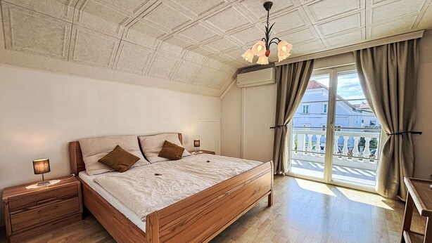 Ein Schlafzimmer mit einem hölzernen Bett, einem Nachttisch mit Lampe, einer Decke mit Dekor und Blick auf einen Balkon durch ein Fenster.