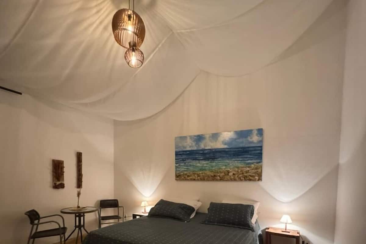 Une chambre à coucher avec un grand lit, un tableau représentant une plage, une table ronde avec des chaises et un luminaire suspendu.