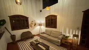 Un salon chaleureux avec un canapé, une table basse et un tapis à rayures. Il y a deux fenêtres avec des cadres en bois, une lampe et une plante sur une table d’appoint.