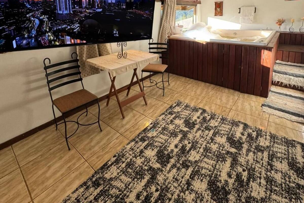 Un salon moderne doté d’un grand téléviseur diffusant une vue sur la ville, d’une table à manger en bois avec des chaises, d’un coin salon confortable avec un canapé et d’un tapis à motifs.