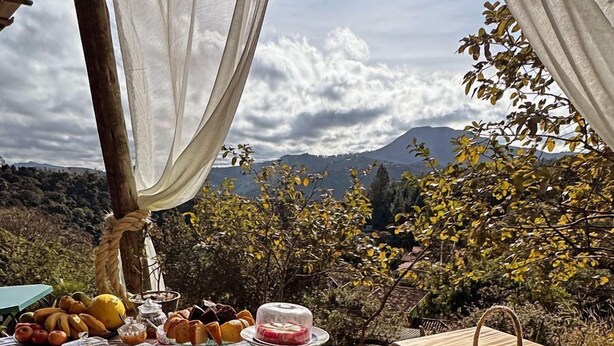 Un espace de restauration en plein air avec différentes sortes de pains et de viennoiseries disposées sur une table en bois, le tout agrémenté d’un magnifique paysage montagneux en arrière-plan.