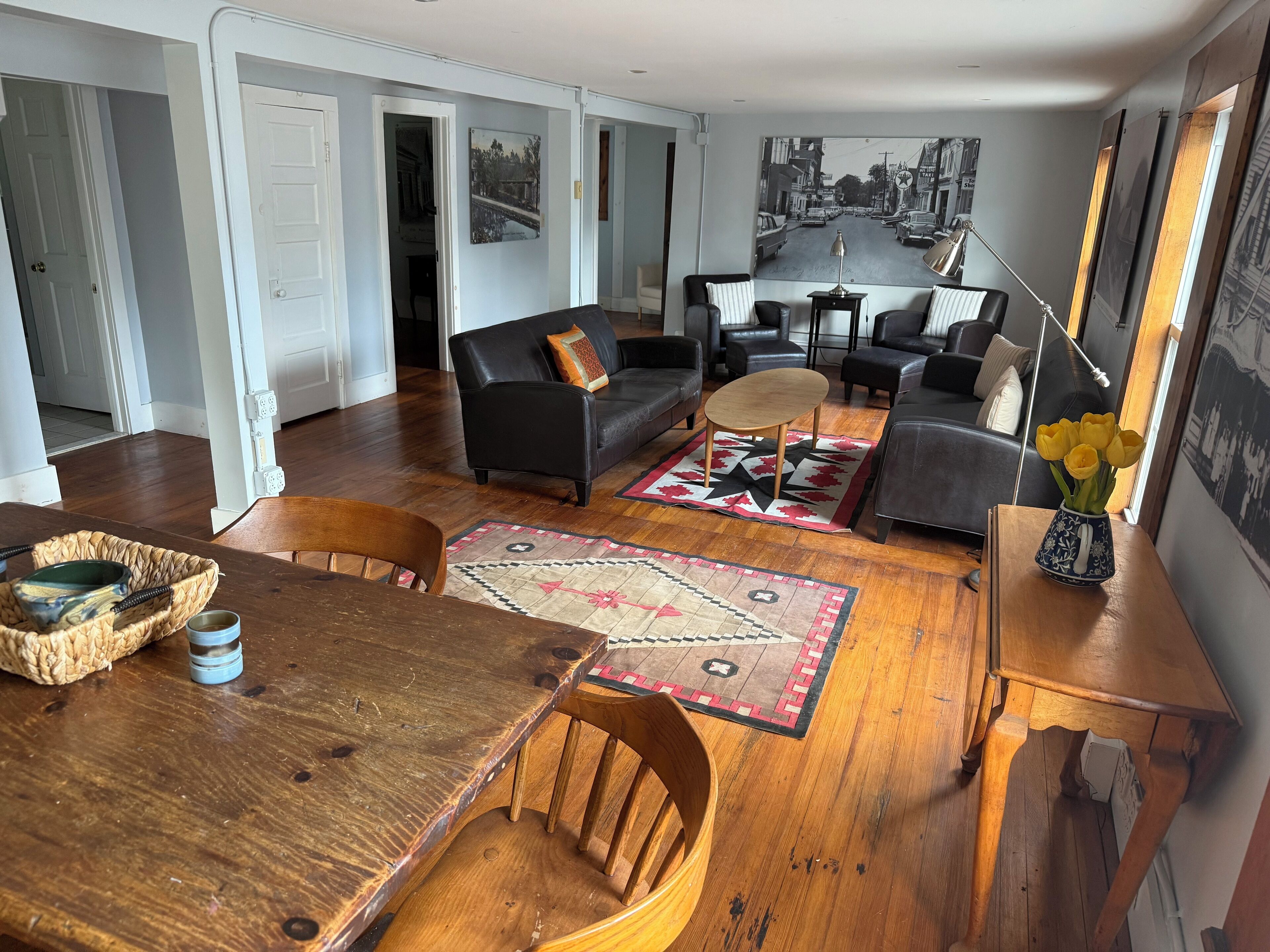 A spacious living area with a wooden dining table, a black leather sofa, and a coffee table. A large window offers a view of a street scene.