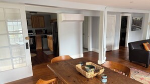 Un coin repas avec une table en bois, des chaises et un panier. On aperçoit, au fond, une cuisine avec des placards en bois et un réfrigérateur.