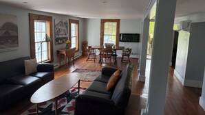 Un salon moderne avec un canapé, une table basse et un coin repas comprenant des chaises et une table en bois. Un téléviseur est fixé au mur et une fenêtre offre une vue sur les arbres.