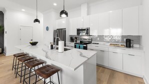 A modern kitchen with white cabinetry, a marble island, bar stools, stainless steel appliances, and pendant lighting.