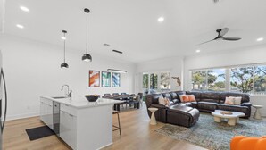 A modern kitchen with a central island, a dining area with a dark leather sofa, and a large window offering a view of the outdoors.
