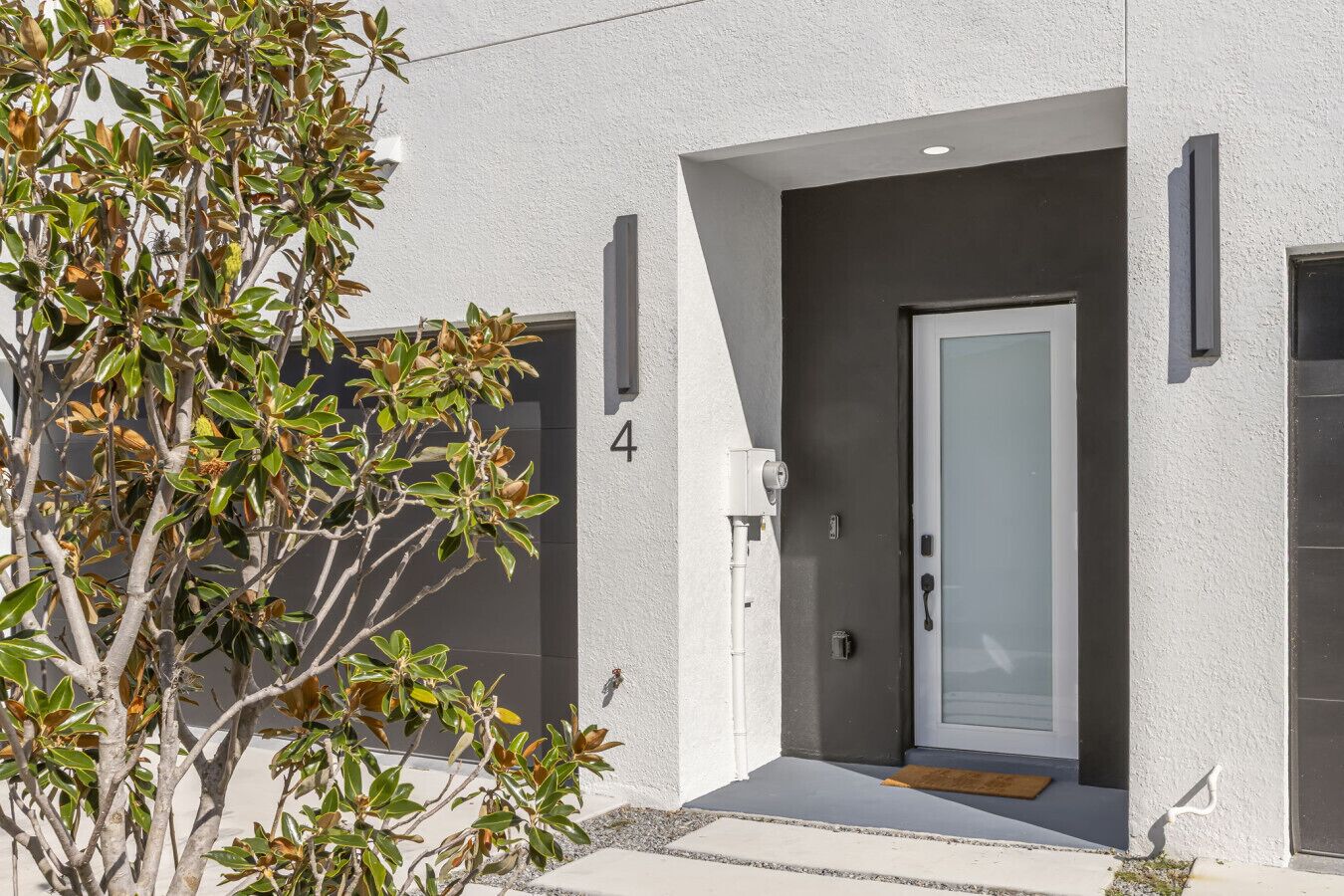A modern building with a white exterior, a numbered door, and a small tree in front.