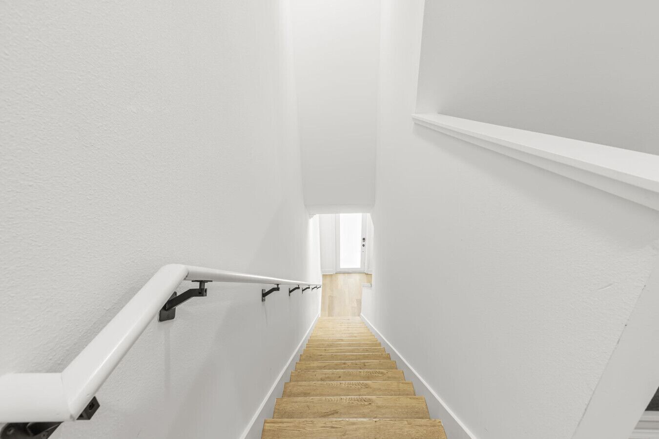 Una escalera estrecha con escalones de madera y paredes blancas, que conduce a una habitación luminosa con ventana.