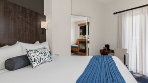 Habitación de hotel con una cama grande, cabecera de madera, mesita de noche con lámpara, una silla y vistas a una chimenea a través de una puerta abierta.