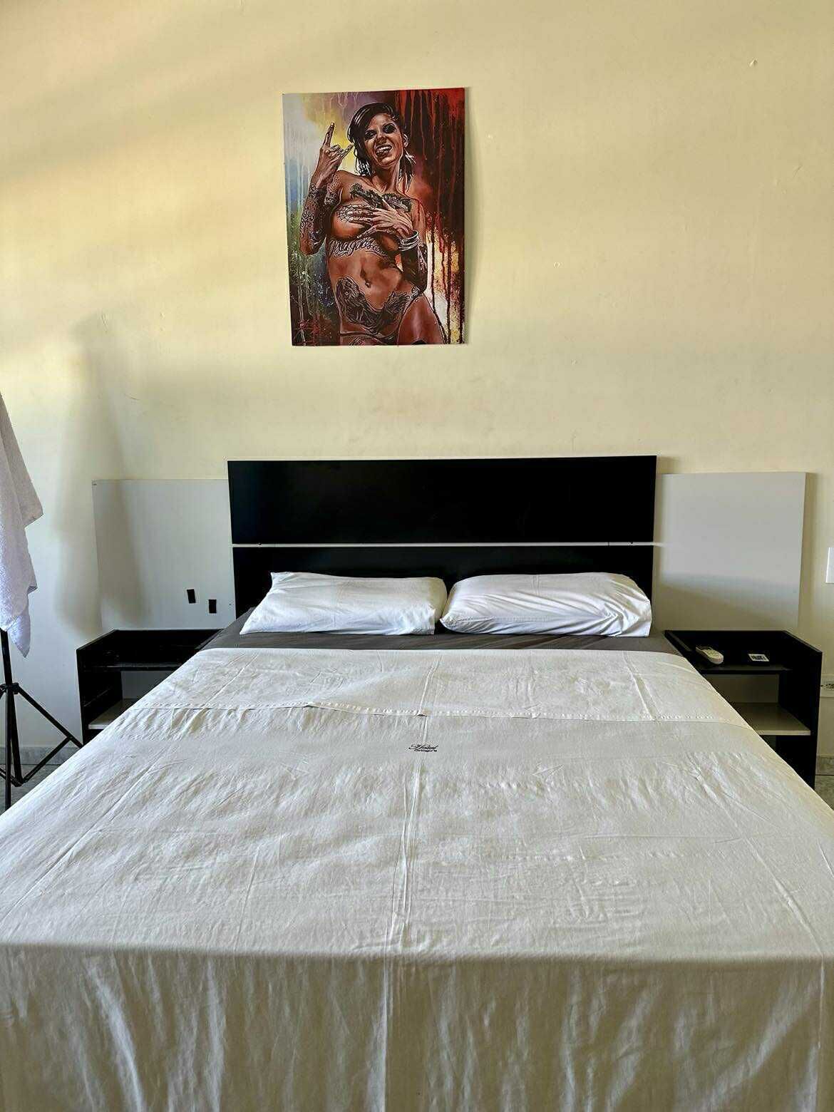 Una habitación de hotel con una cama grande, ropa de cama blanca, cabecero negro, dos mesitas de noche y un cuadro en la pared.
