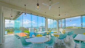A modern dining area with round tables and chairs, large windows offering a city view, and hanging pendant lights. - Wonmi Residency (Galle)