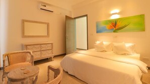 A hotel room with a bed, a chair, a small table, a dresser, a painting, and an air conditioning unit. - Wonmi Residency (Galle)