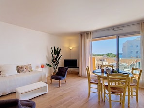 A living room with a sofa, a TV, a dining table with chairs, a plant, and a balcony with a view.