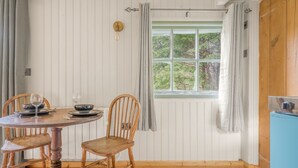 A dining area with a round wooden table, two wooden chairs, a window with a view of greenery, and a blue cabinet. - Bluebell Lodge, Lake District Shepherd's Hut for 2 with hot tub (Coniston)