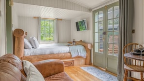 A cozy room with a bed, a wooden bench, a table with a chair, and a large window with curtains. - Bluebell Lodge, Lake District Shepherd's Hut for 2 with hot tub (Coniston)