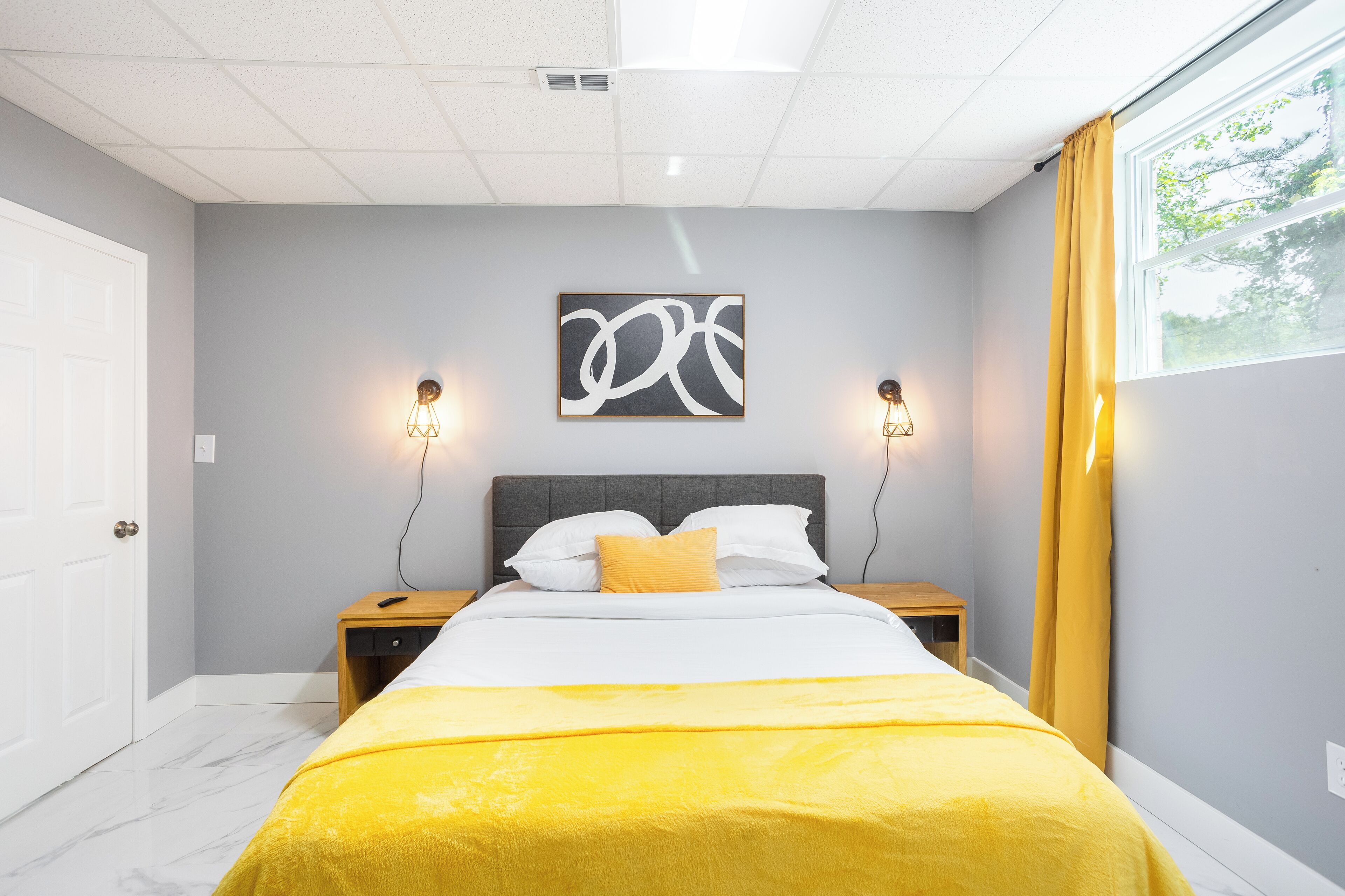 A bedroom with a bed, a bedside table, a nightstand, a wall-mounted artwork, and a window with curtains.