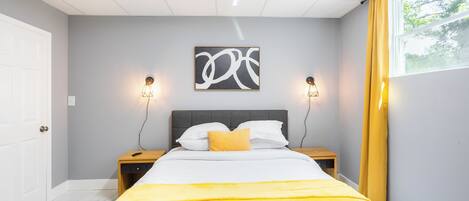 A bedroom with a bed, a bedside table, a nightstand, a wall-mounted artwork, and a window with curtains.