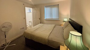 Une chambre à coucher avec un lit, un ventilateur sur pied, une fenêtre avec des stores et une table de chevet avec une lampe.