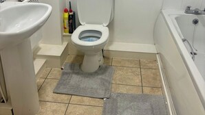 Une salle de bain avec des toilettes, un lavabo et une baignoire. Il y a deux tapis gris sur le sol.