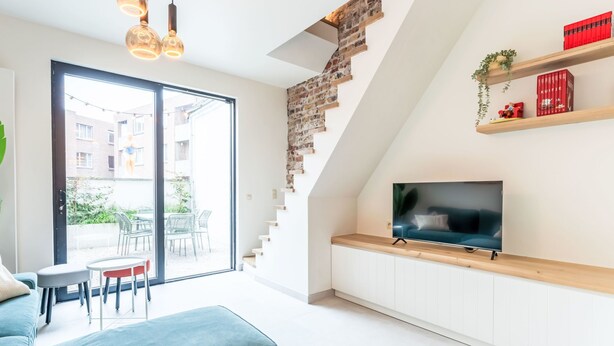 Een moderne woonkamer met een grote televisie, een trap met onbehandelde bakstenen en uitzicht op buiten.