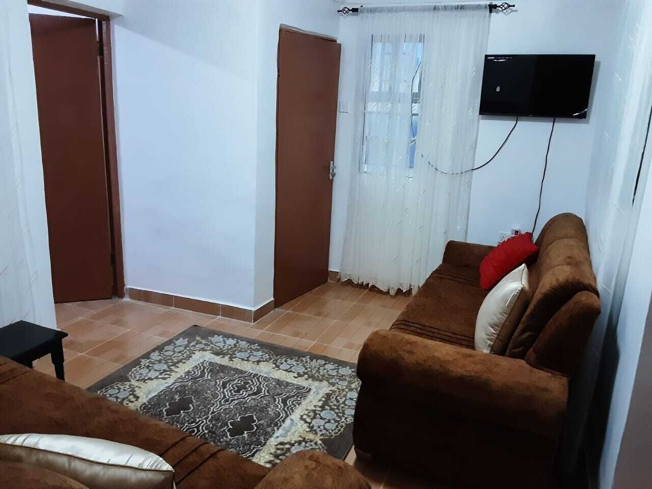 A living room with a brown sofa, a small table, a patterned rug, a television mounted on the wall, and a door with a curtain.