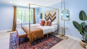 Een slaapkamer met een hemelbed, een bankje, een kamerkleed met dessin, een spiegel en een potplant.