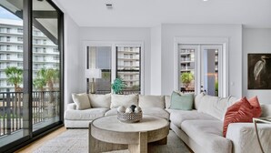 A modern living room with a large sectional sofa, a round coffee table, and a view of a high-rise building through large windows. - Arcadia Sands: Private Beach, Ocean Views, Pool & Hot tub, Game Room, Bunk Rooms (Miramar Beach)