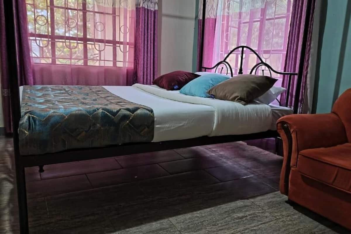 Una cama individual con una colcha estampada, una silla roja y una ventana con cortinas moradas.