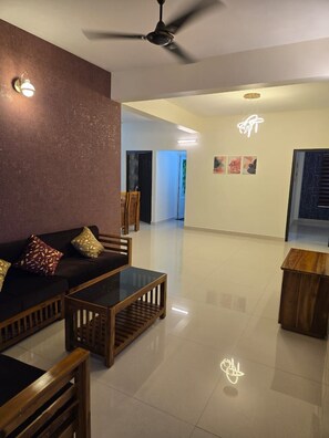 A spacious living room with a dark-colored sofa set, a wooden coffee table, and a ceiling fan. - Centur Apartments (Changanassery)