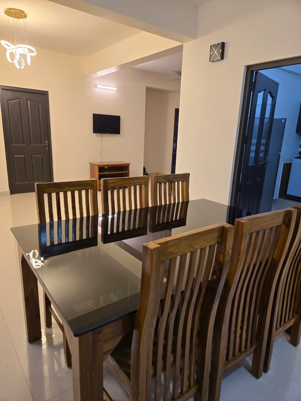 A dining area with a glass table and wooden chairs, a television on the wall, and a door leading to another room.