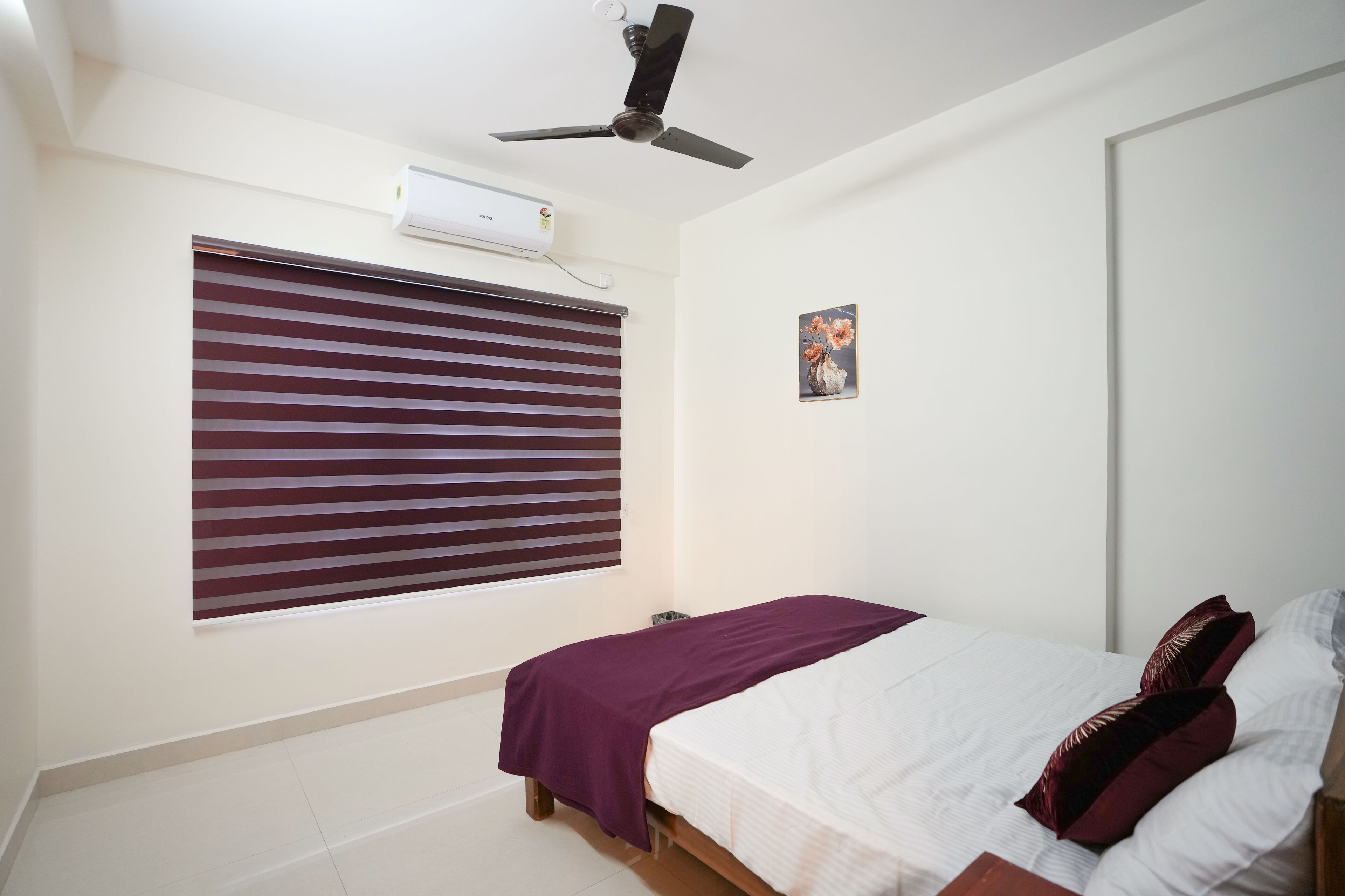A bedroom with a bed, a ceiling fan, an air conditioning unit, a window with blinds, and a small picture on the wall.