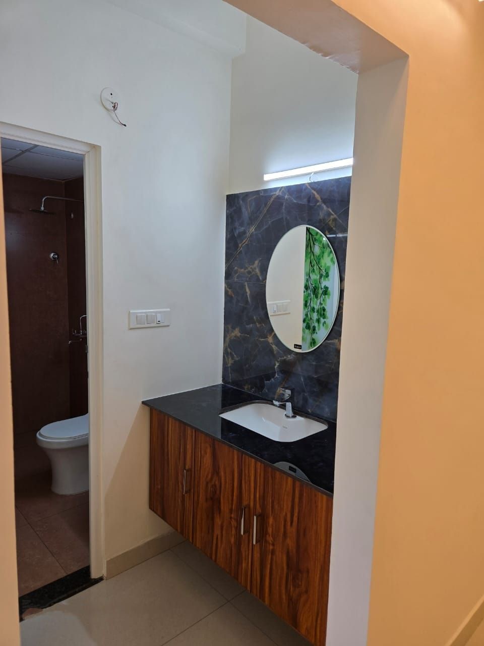 Un baño con inodoro blanco, lavamanos sobre encimera negra, armarios de madera y salpicadero de mármol con estampado de hojas verdes.