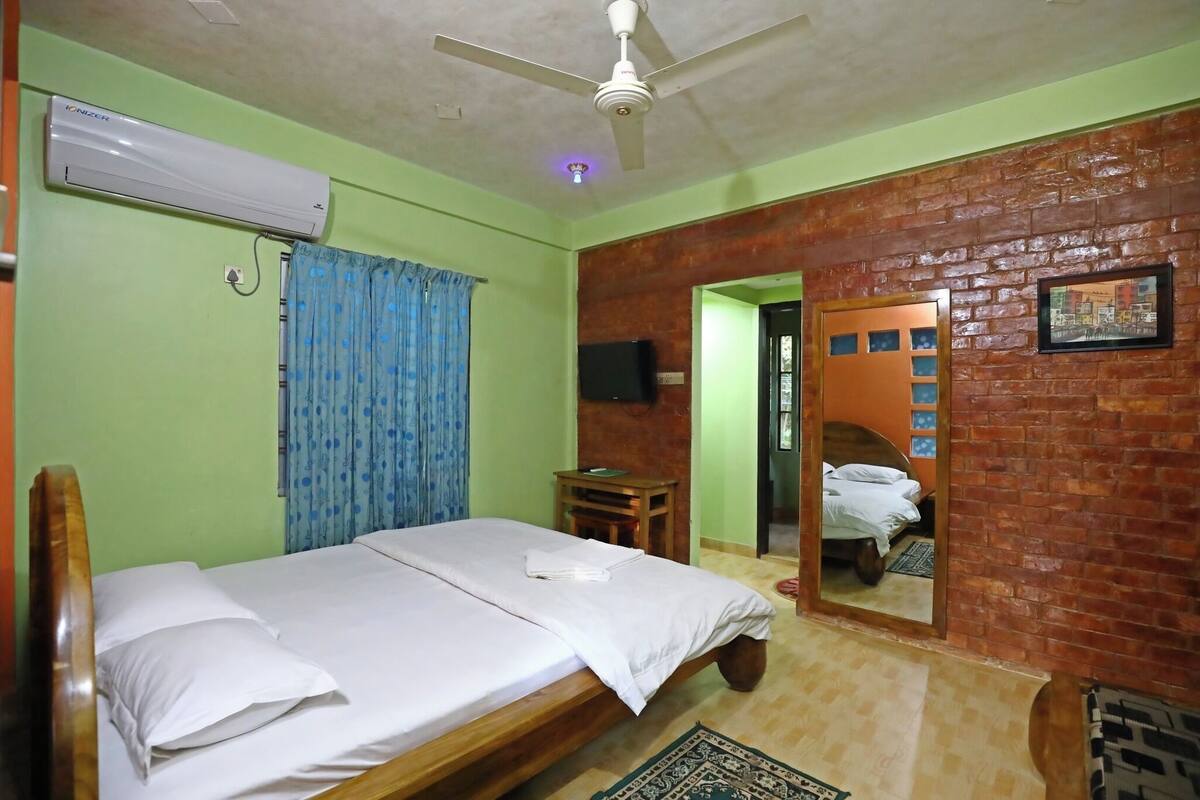 Une chambre comprenant un lit, un climatiseur, un ventilateur de plafond, une table en bois, un téléviseur et un miroir qui reflète le lit.