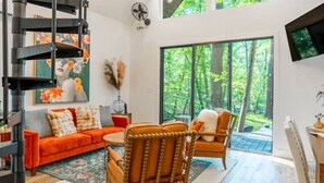A modern living room with a wooden floor, a sofa, two chairs, a TV, and a staircase. - Romantic Cabin | Hot Tub, Soaking Tubs & Fire Pit (Bullard)