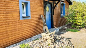 Una baita in legno con una porta e delle finestre blu, circondata da un vialetto sterrato e vegetazione.
