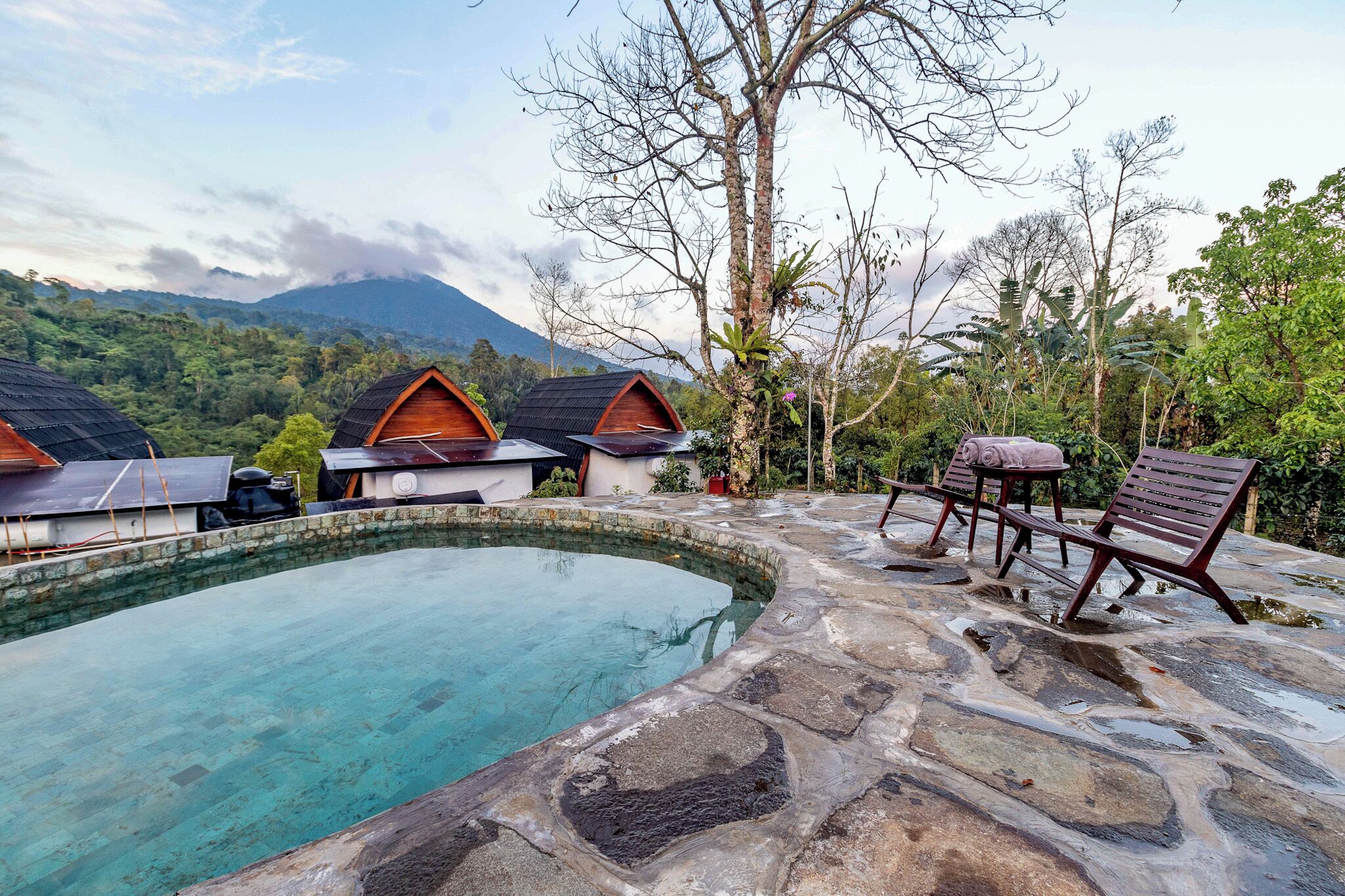 Zona de piscina con suelo de piedra, sillones de madera y vistas a montañas al fondo.