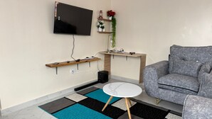 A modern living room with a grey sofa, a flat-screen TV mounted on the wall, wooden shelves, a small table, and a patterned rug.