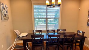 Un comedor con una mesa y sillas de madera oscura, un techo alto con una araña, y una ventana con persianas.