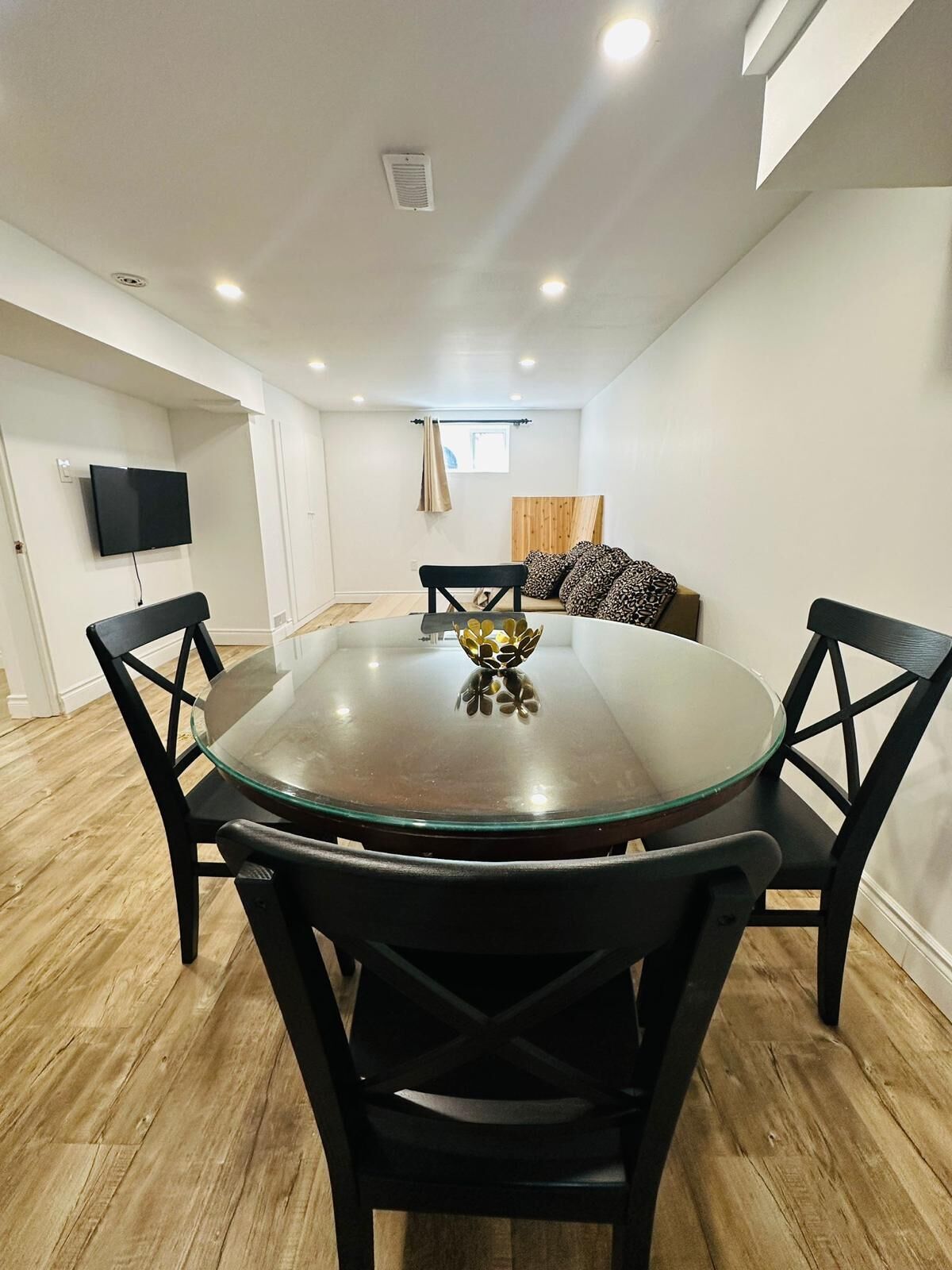 Un comedor con una mesa redonda de vidrio, sillas negras, un cuenco decorativo, un televisor en la pared y una ventana con cortinas.