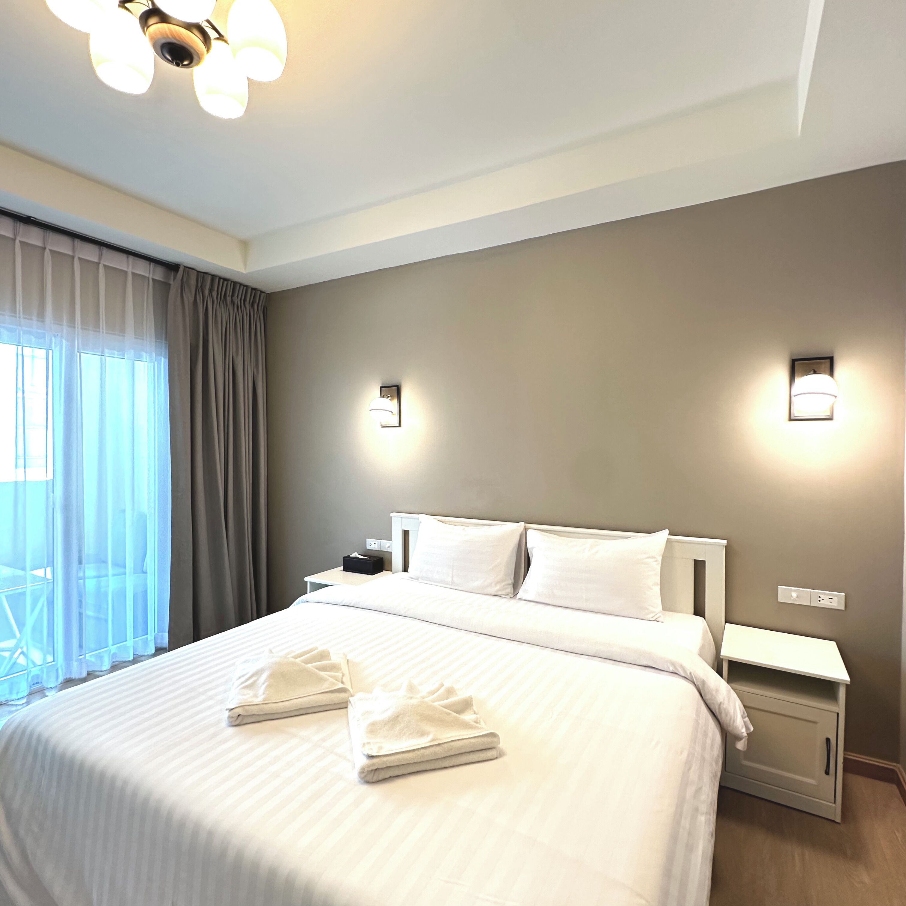 Quarto de hotel com uma cama grande, duas mesas de cabeceira, um lustre no teto e uma janela com cortinas.