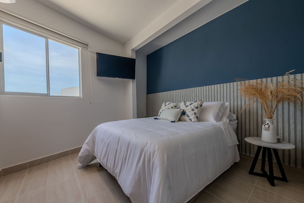 A bedroom with a bed, a TV mounted on the wall, a window, and a vase with dried plants on a small table.