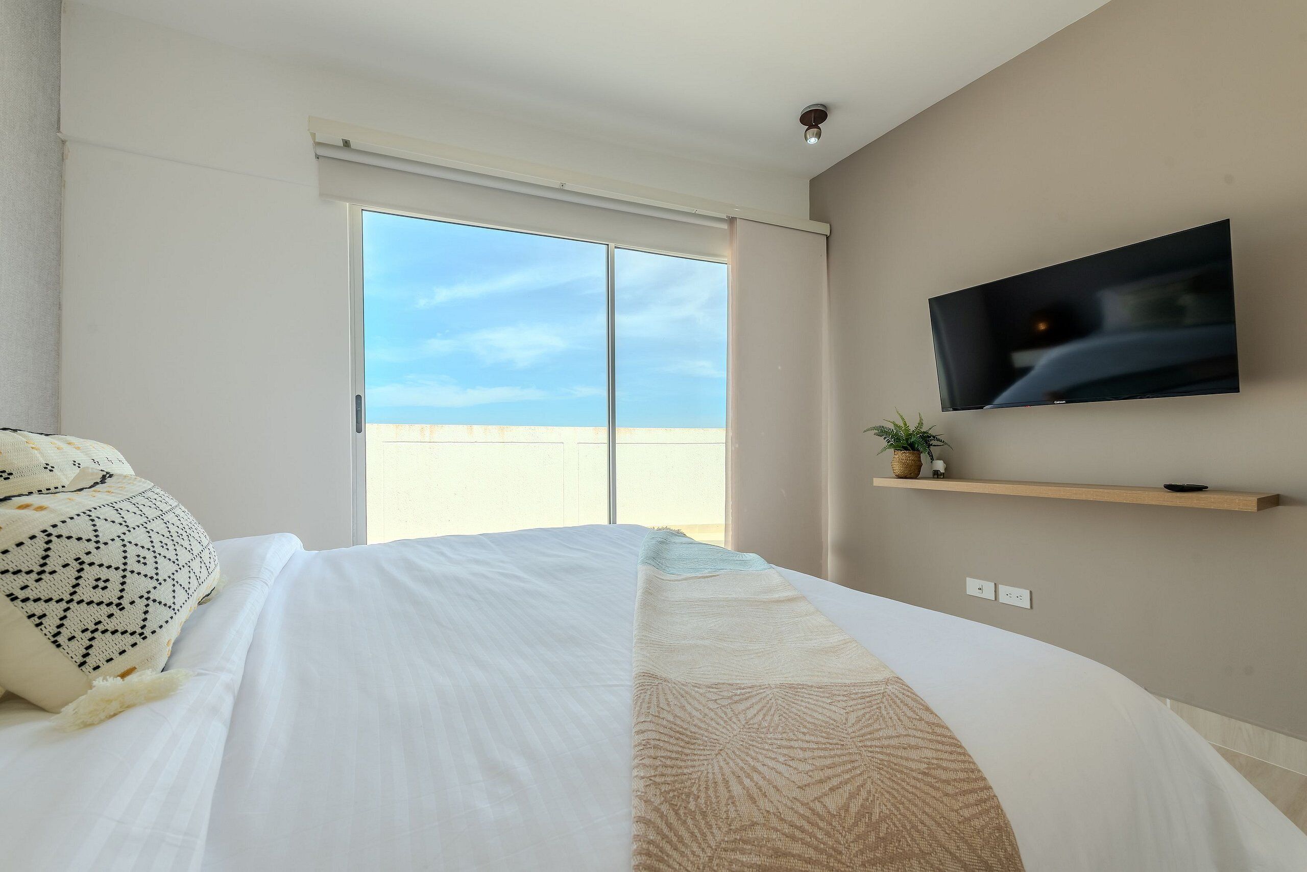 Ein Schlafzimmer mit Bett, einem an der Wand befestigten Fernseher, einer Schiebeglas-Tür zum Balkon und einem kleinen Regal mit einer Pflanze.