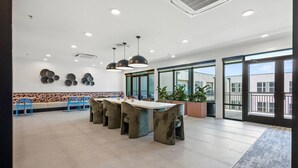 A spacious indoor area with a long table and chairs, large windows, and potted plants. - Trendy Bishop Arts | Game Room + Gym + Yoga (Dallas)