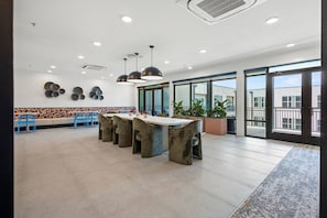 Un espacio interior amplio con una larga mesa y sillas, ventanas grandes y plantas en macetas.