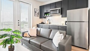 A modern kitchen with dark cabinets, a stainless steel refrigerator, and a microwave. There is a grey leather sofa with white cushions and a potted plant.