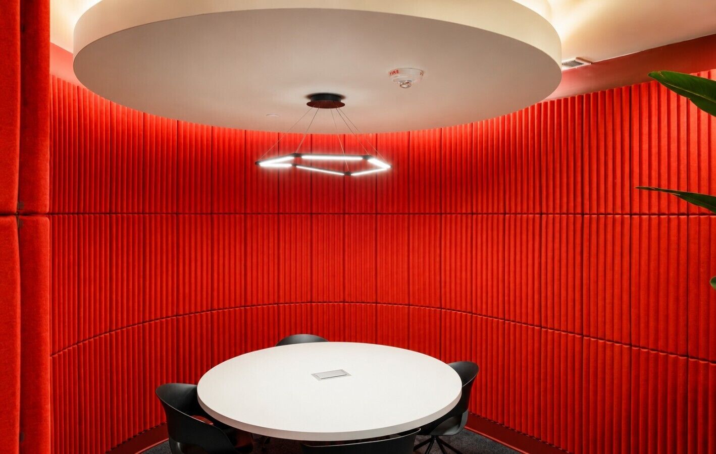 A circular conference table with a white top, surrounded by black chairs, in a room with red vertical paneling and a ceiling light fixture.