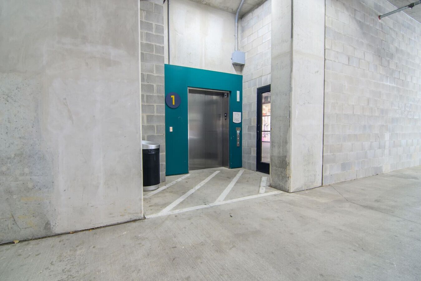 An elevator in a building with a teal door, a trash bin, and a concrete floor.