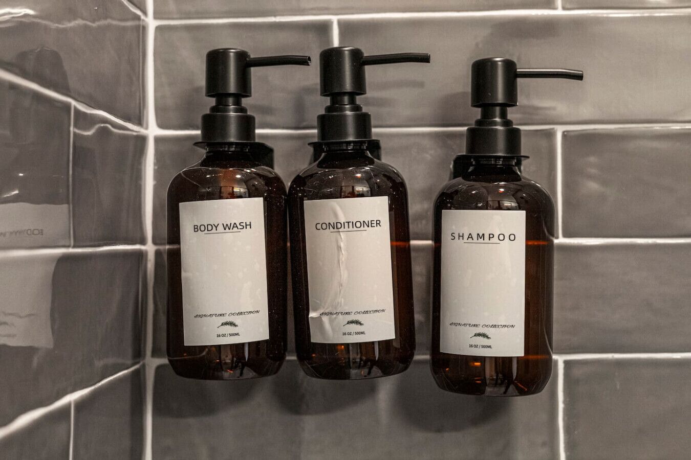 Three bottles of body wash, conditioner, and shampoo with pump dispensers, mounted on a tiled wall.