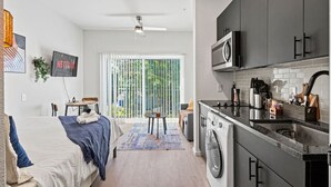 A modern apartment with a kitchenette, a bed with a blue blanket, a TV mounted on the wall, and a view of the outdoors through a sliding glass door.