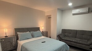 A bedroom with a bed, bedside tables, a grey sofa, and an air conditioning unit. - Bienes House private property in Brandon (Brandon)